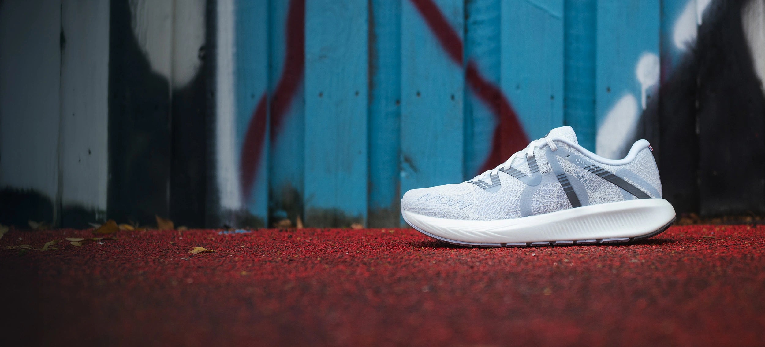 Seitenansicht eines weißen MOVV Laufschuhs, der auf einer strukturierten roten Oberfläche, wahrscheinlich einer Laufbahn oder einem Gummiboden, platziert ist. Der Hintergrund zeigt eine blaue Holzwand mit Graffiti-ähnlicher Bemalung in Rot, Weiß und Schwarz. Das Schuhdesign umfasst subtile geometrische Muster und halbtransparente Überlagerungen.