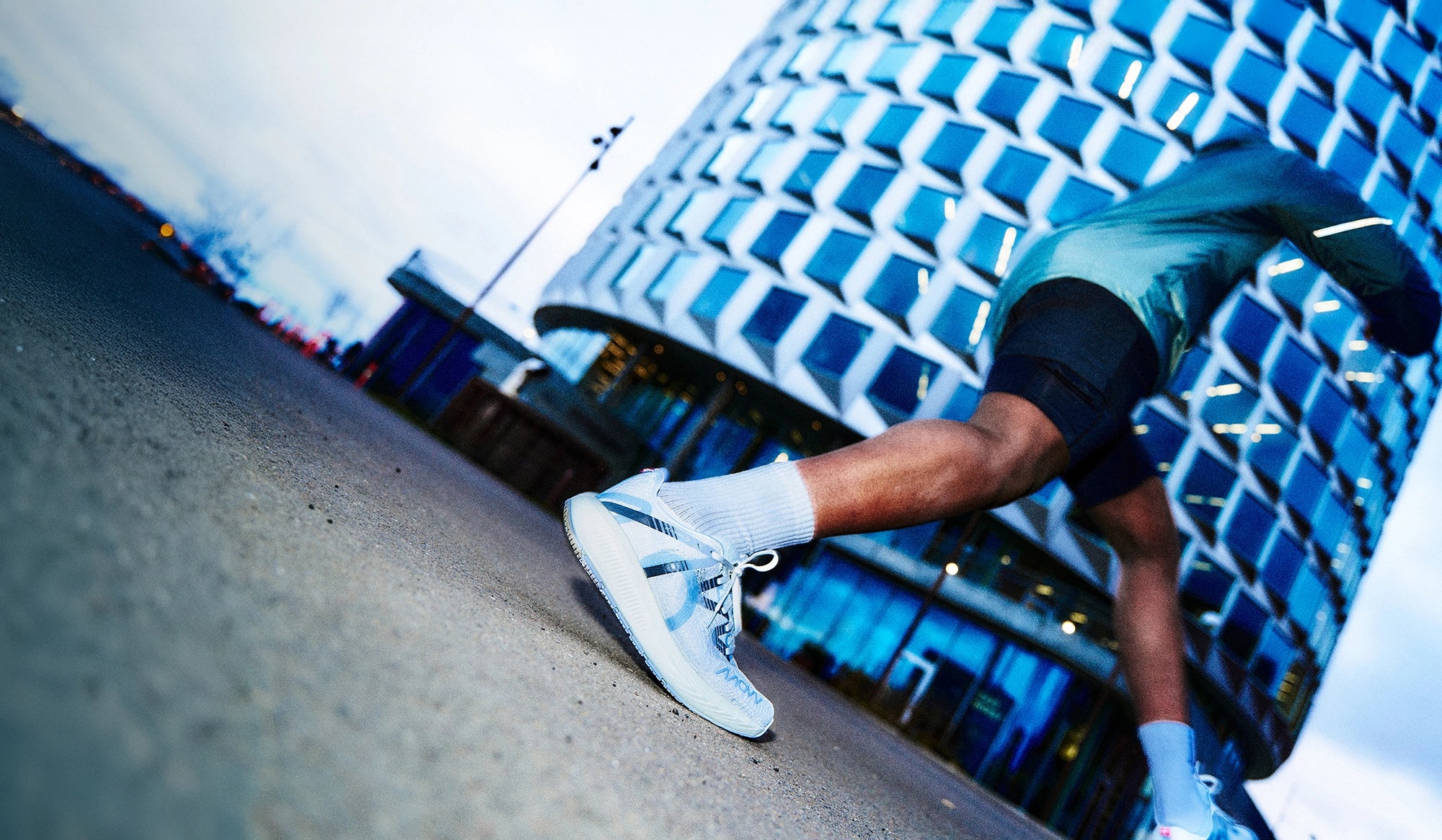 Läufer in der Mitte des Schritts auf einer städtischen Straße, trägt hellblaue MOVV Laufschuhe und weiße Socken. Im Hintergrund befindet sich ein modernes, geometrisches Glasgebäude mit blauem Schimmer, aufgenommen in der Dämmerung. Die Bewegungsunschärfe betont Geschwindigkeit und Energie.
