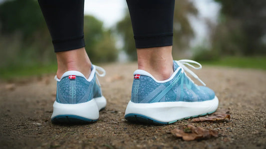 Close-up of blue running shoes with white soles on a dirt trail, worn by person in black leggings