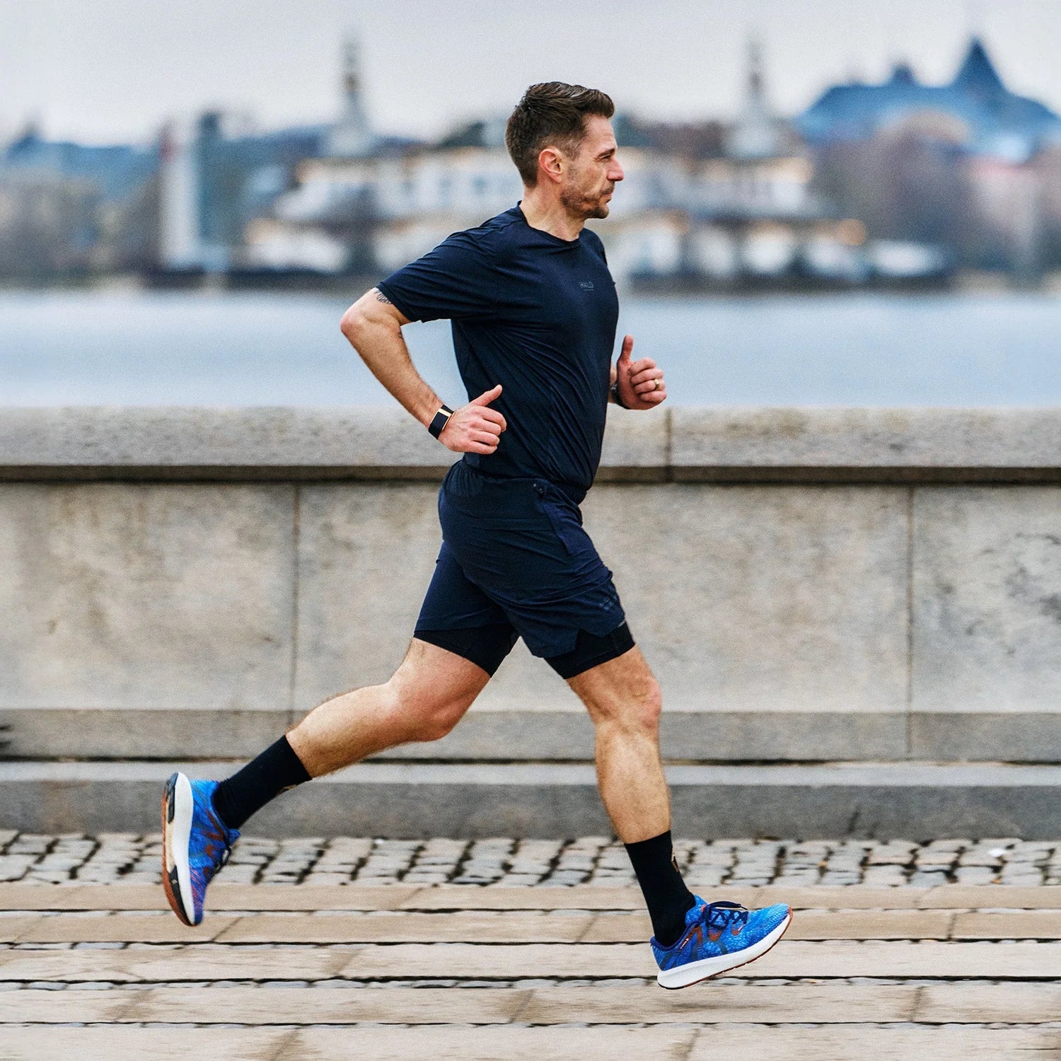Man running along a waterfront promenade wearing a navy blue athletic outfit, blue MOVV Svandinavian running shoes, and a fitness tracker, with a blurred cityscape in the background.