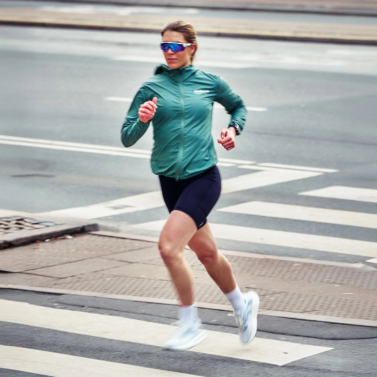 Woman running across a city street in athletic gear, wearing a green jacket, black shorts, white MOVV Scandinavian running shoes, and blue-tinted sports sunglasses.
