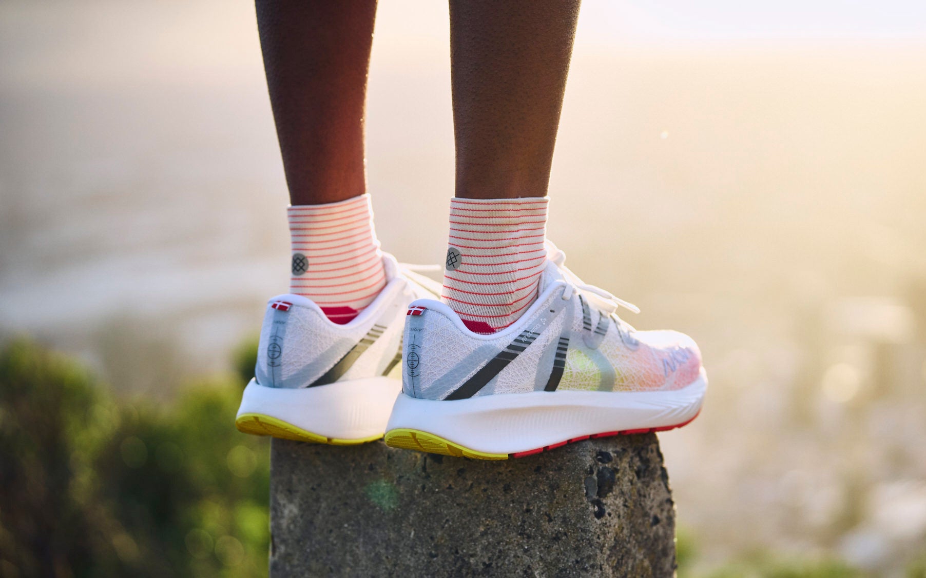 Close-up of a runner's feet wearing white MOVV running shoes and striped socks, standing on a rock overlook at sunset.