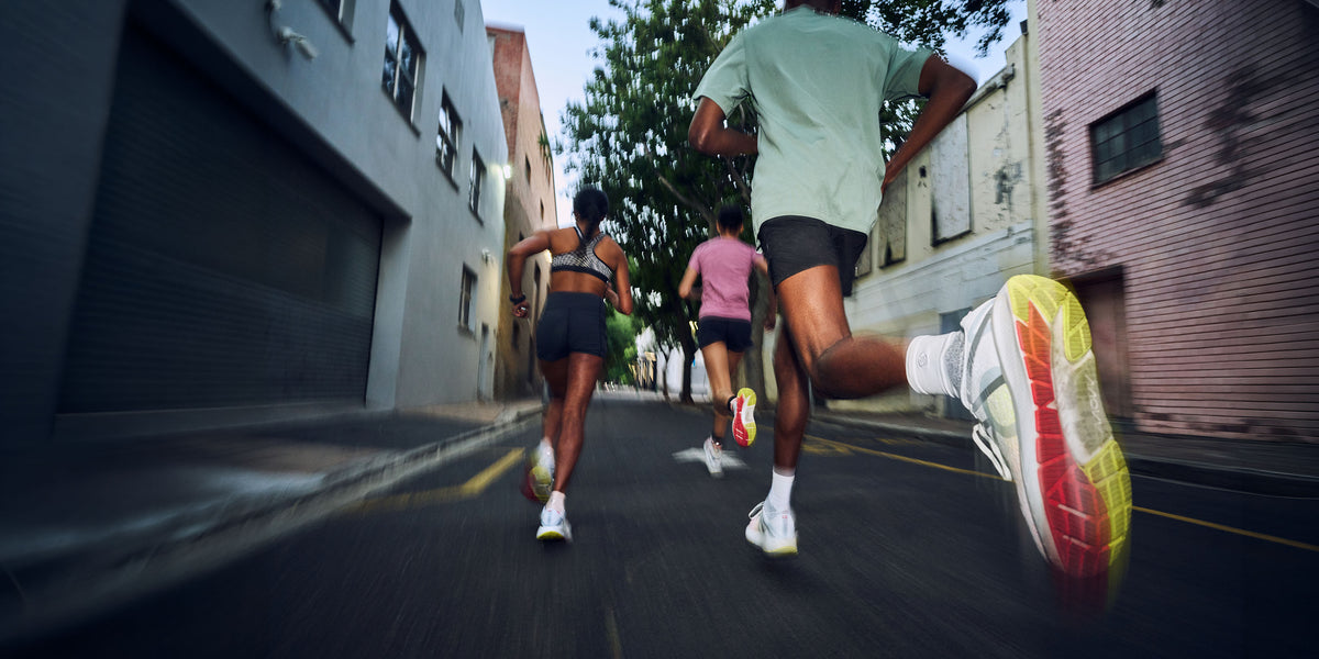 Three runners from behind on an urban street, with the foreground runner's legs and colorful running shoe prominently shown in motion.