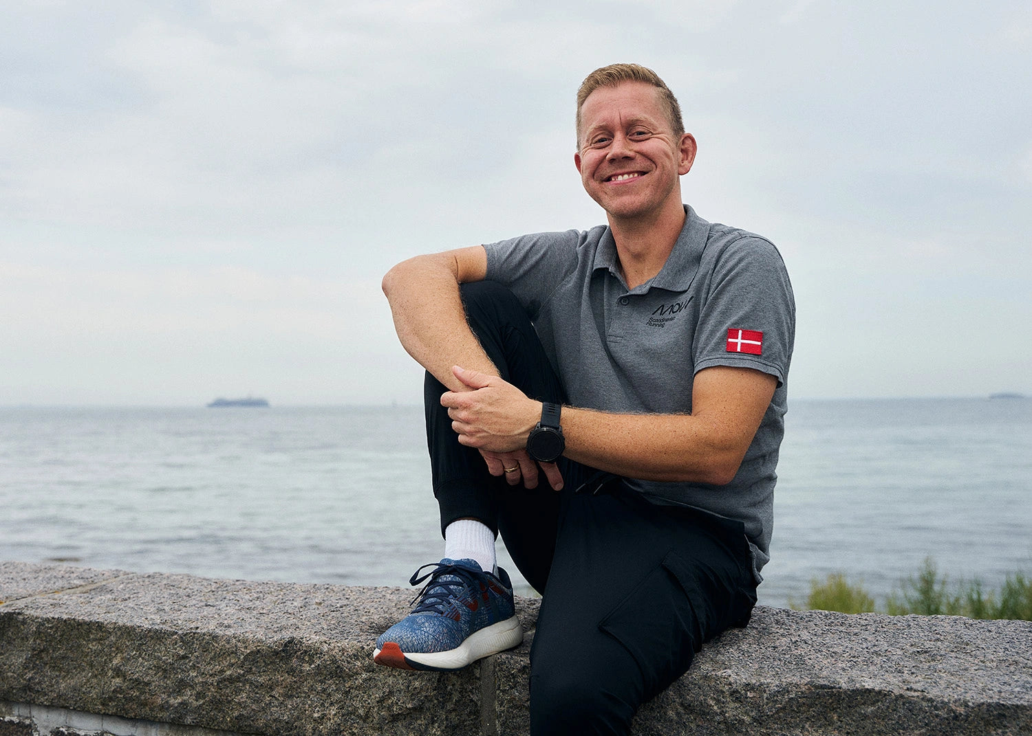 Man sitting on a ledge by the water wearing a gray polo shirt with a MOVV logo.