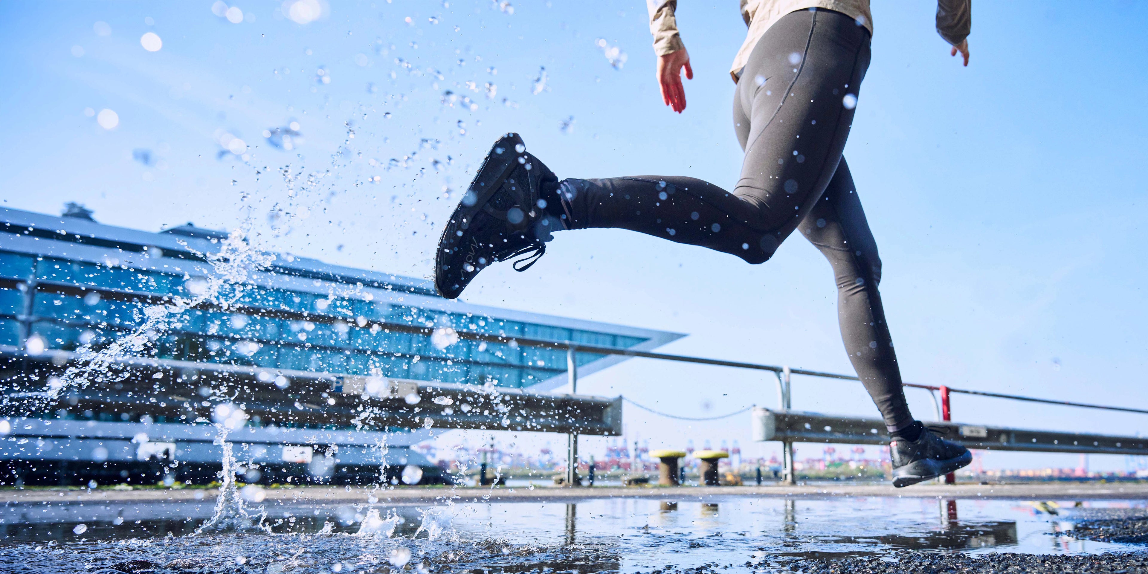 A person running outdoors in black athletic leggings and black MOVV running shoes, with water splashing around their leg in a wet urban setting under a blue sky.